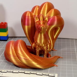 May include: A 3D printed figurine of a nine-tailed fox creature with a metallic gradient finish shifting between orange, gold, and pink. The fox sits upright with multiple curved tails fanned out behind and around it, positioned near a ruler.