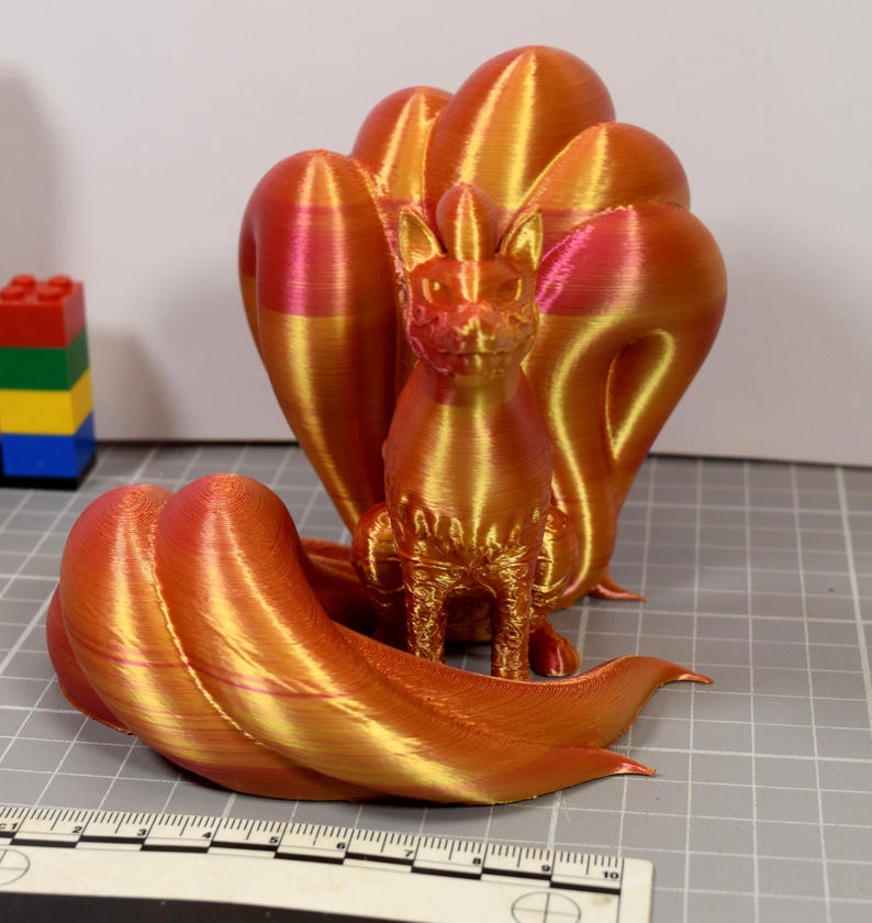 May include: A 3D printed figurine of a nine-tailed fox creature with a metallic gradient finish shifting between orange, gold, and pink. The fox sits upright with multiple curved tails fanned out behind and around it, positioned near a ruler.