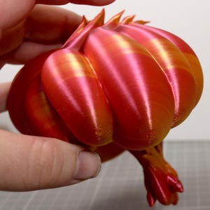 May include: A 3D printed object with a rounded, segmented shape featuring a gradient of metallic red and orange colors. The surface displays visible concentric circular layer lines from the printing process, with pointed protrusions at the top.