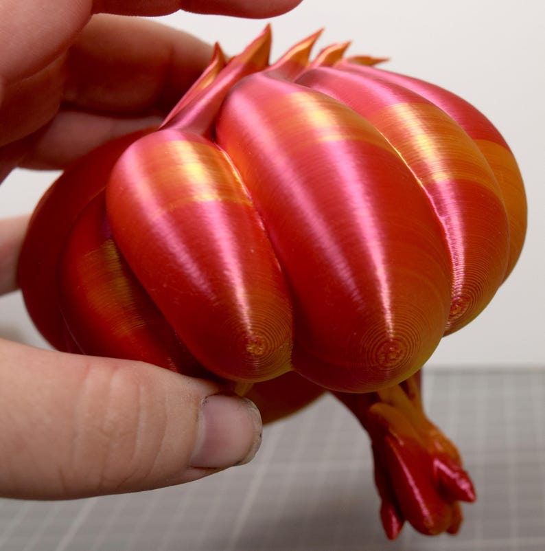 May include: A 3D printed object with a rounded, segmented shape featuring a gradient of metallic red and orange colors. The surface displays visible concentric circular layer lines from the printing process, with pointed protrusions at the top.