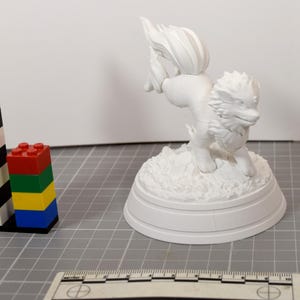May include: A white, unpainted 3D printed figurine of a four-legged creature with a bushy tail and spiky fur, standing on a textured circular base. A ruler at the bottom shows a scale of 10 centimeters, and a stack of colorful plastic bricks is nearby.