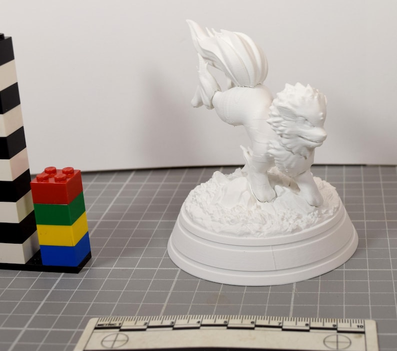 May include: A white, unpainted 3D printed figurine of a four-legged creature with a bushy tail and spiky fur, standing on a textured circular base. A ruler at the bottom shows a scale of 10 centimeters, and a stack of colorful plastic bricks is nearby.
