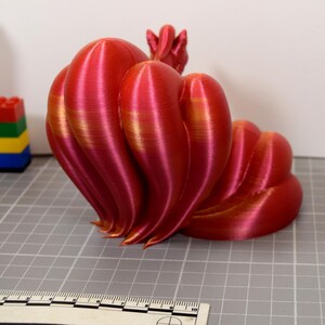 May include: A 3D printed nine-tailed fox figurine featuring a gradient of metallic red and gold colors. The figure has multiple curved, flowing tails and is positioned next to a ruler showing a scale of 10 centimeters for size reference.