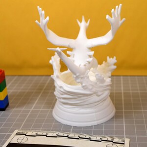 May include: A white 3D printed figurine of a creature with large wings and a long tail, emerging from a swirling water-like base. A ruler is visible in the foreground, showing a scale in centimeters, and a small stack of colorful plastic blocks.