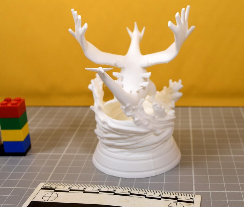 May include: A white 3D printed figurine of a creature with large wings and a long tail, emerging from a swirling water-like base. A ruler is visible in the foreground, showing a scale in centimeters, and a small stack of colorful plastic blocks.