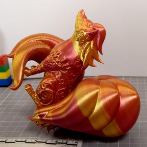 May include: A 3D printed fox figurine with a gradient of metallic red, orange, and gold colors. The fox features intricate swirling patterns on its body and a large, segmented tail. A ruler is visible at the base, showing a scale in inches.