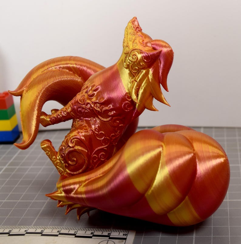 May include: A 3D printed fox figurine with a gradient of metallic red, orange, and gold colors. The fox features intricate swirling patterns on its body and a large, segmented tail. A ruler is visible at the base, showing a scale in inches.