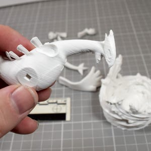May include: A white 3D printed figurine kit featuring a creature head with a long neck, separate limb components, and a textured circular base. The pieces are unpainted and show visible layer lines from the printing process on a grid surface.