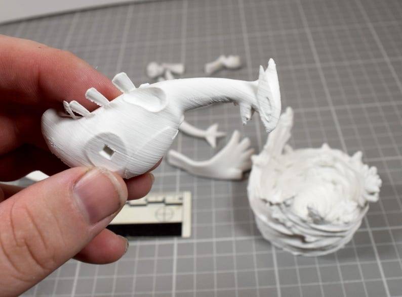May include: A white 3D printed figurine kit featuring a creature head with a long neck, separate limb components, and a textured circular base. The pieces are unpainted and show visible layer lines from the printing process on a grid surface.