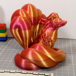 May include: A 3D printed fox figurine with nine tails, featuring a gradient of metallic red, orange, and gold colors. The surface displays visible horizontal layer lines from the printing process. A ruler is visible at the base for scale.