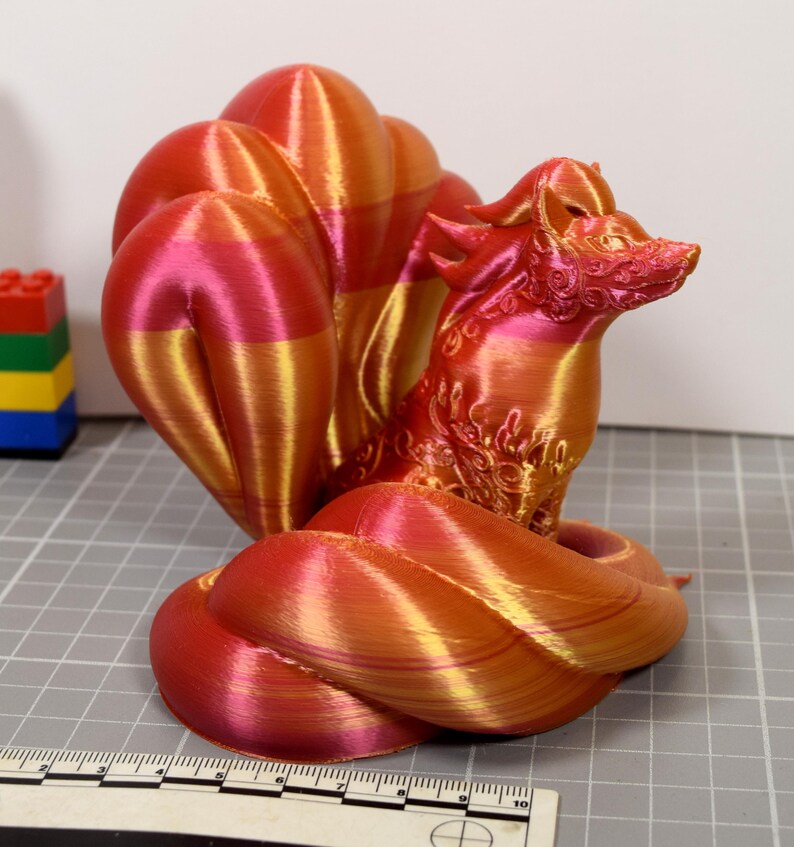 May include: A 3D printed fox figurine with nine tails, featuring a gradient of metallic red, orange, and gold colors. The surface displays visible horizontal layer lines from the printing process. A ruler is visible at the base for scale.