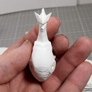 May include: A small, white, 3D-printed figurine featuring a creature with a long, slender neck, pointed ears, and a smooth, rounded body. The surface displays fine horizontal layer lines characteristic of additive manufacturing processes.