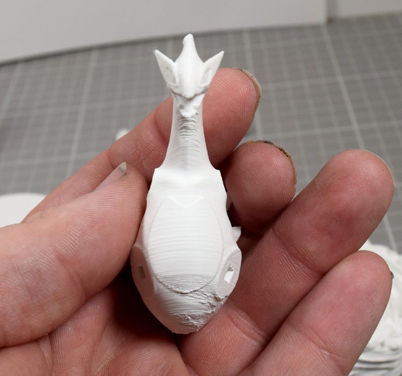 May include: A small, white, 3D-printed figurine featuring a creature with a long, slender neck, pointed ears, and a smooth, rounded body. The surface displays fine horizontal layer lines characteristic of additive manufacturing processes.