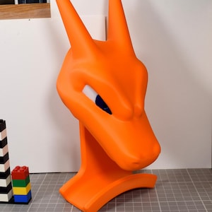 May include: A bright orange dragon-like creature head bust featuring two tall pointed horns and narrow blue eyes. The sculpture is positioned on a gray grid surface next to a white ruler showing measurements in inches and a small stack of colorful plastic bricks.
