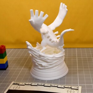 May include: A white 3D printed figurine of a creature with large wings and a long tail, positioned atop a sculpted base resembling swirling water. A ruler is visible at the bottom, showing the base is approximately 3.5 inches in diameter.