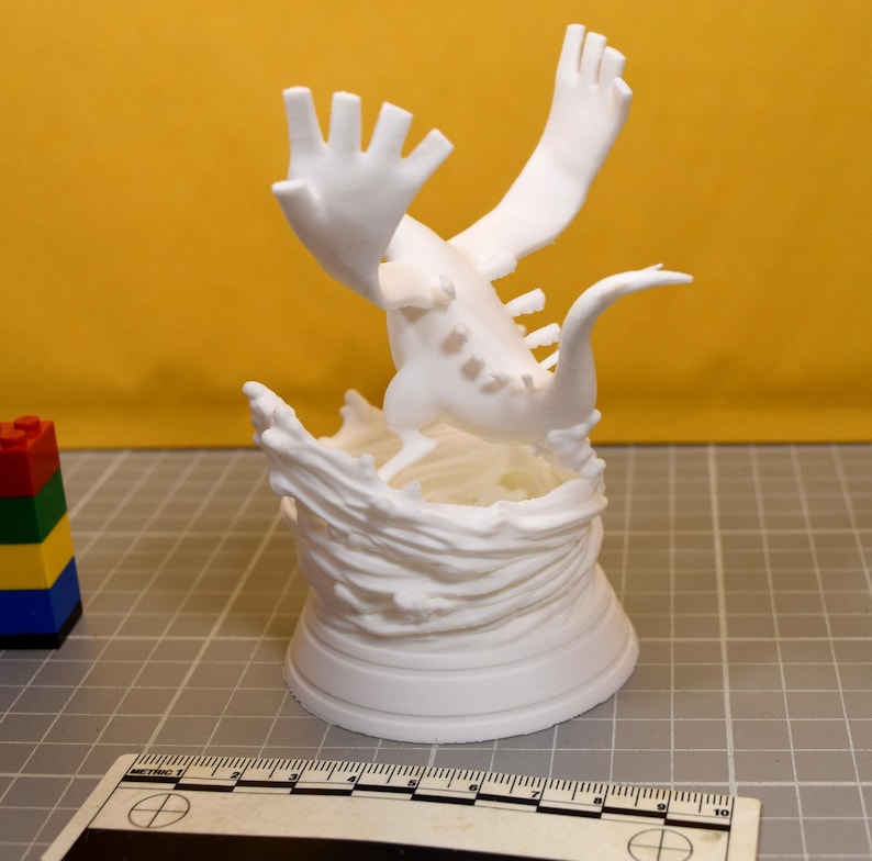 May include: A white 3D printed figurine of a creature with large wings and a long tail, positioned atop a sculpted base resembling swirling water. A ruler is visible at the bottom, showing the base is approximately 3.5 inches in diameter.