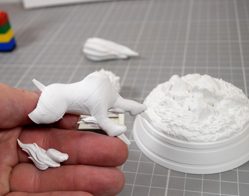 May include: A white unpainted 3D printed miniature of a four-legged creature in a dynamic pose, accompanied by a separate small flame-shaped piece and a circular base with a textured rocky surface, all crafted from a matte white resin material.