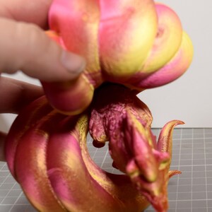 May include: A multi-jointed articulated dragon figurine featuring a vibrant gradient of metallic pink and gold colors. The creature has a detailed head, a textured body, and a long, segmented tail that curves around. A ruler is visible at the base.