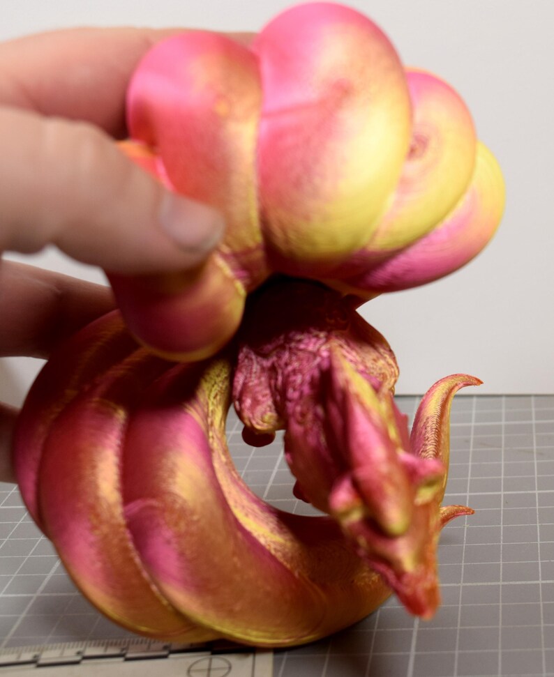 May include: A multi-jointed articulated dragon figurine featuring a vibrant gradient of metallic pink and gold colors. The creature has a detailed head, a textured body, and a long, segmented tail that curves around. A ruler is visible at the base.