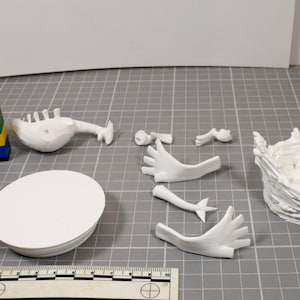 May include: A collection of white 3D printed model parts including a circular base, a torso, two hands, two arms, and a textured circular stand. A black and white striped tower and a small stack of colorful plastic bricks are visible for scale.