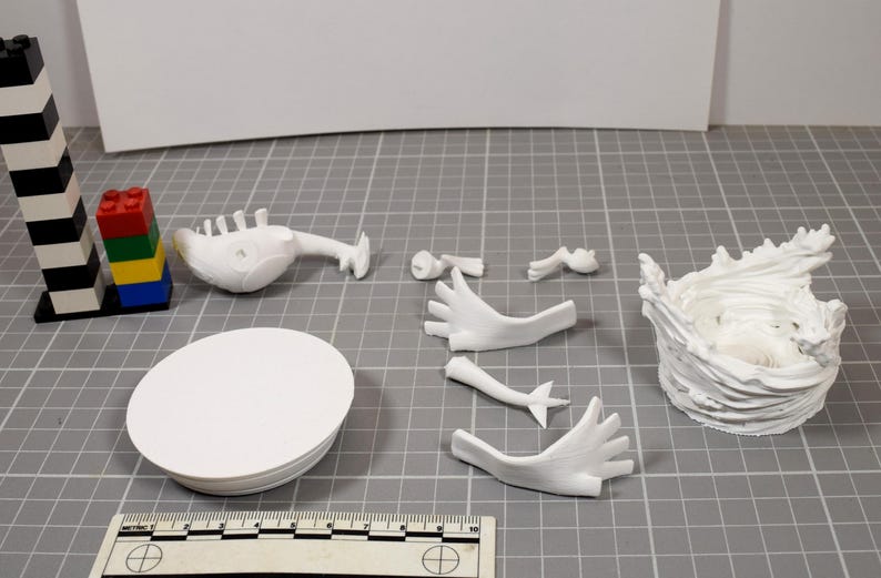 May include: A collection of white 3D printed model parts including a circular base, a torso, two hands, two arms, and a textured circular stand. A black and white striped tower and a small stack of colorful plastic bricks are visible for scale.