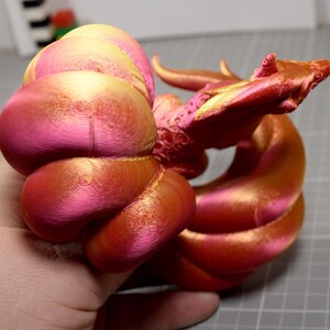May include: A 3D printed dragon figurine featuring a gradient of metallic pink and gold colors. The creature has a coiled body, a detailed head with horns, and a textured surface showing concentric layer lines from the printing process.