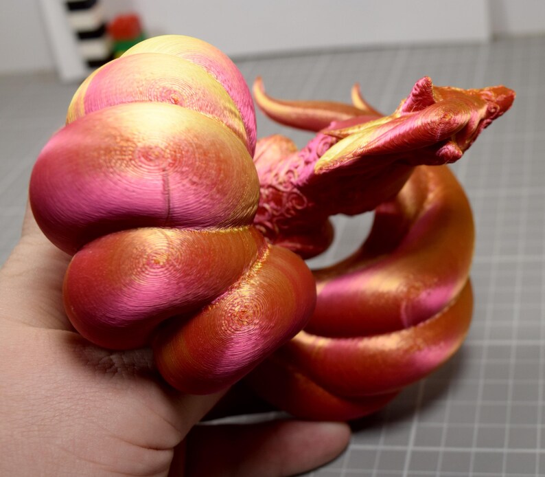 May include: A 3D printed dragon figurine featuring a gradient of metallic pink and gold colors. The creature has a coiled body, a detailed head with horns, and a textured surface showing concentric layer lines from the printing process.