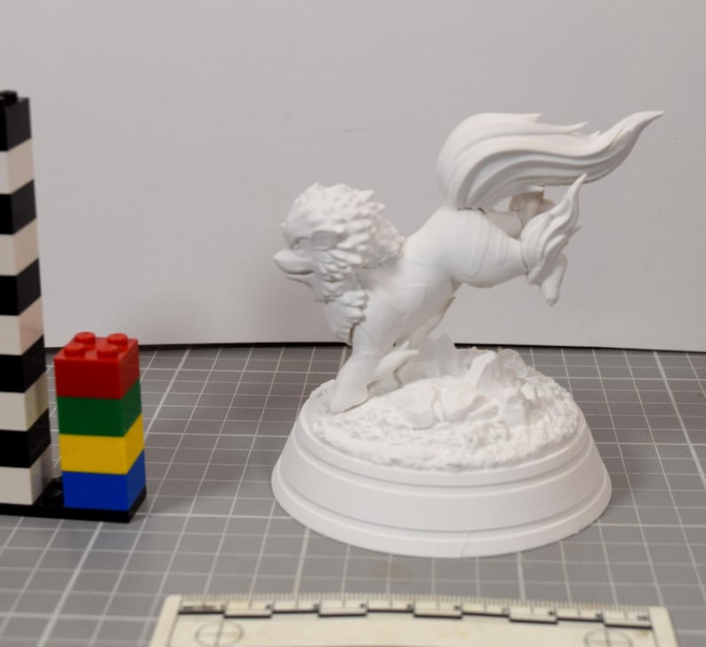 May include: A white, unpainted 3D printed figurine of a mythical creature with a large, flowing tail and a mane, standing on a textured base. A black and white striped tower and a stack of red, green, yellow, and blue plastic bricks are nearby.