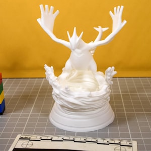 May include: A white 3D printed figurine of a mythical creature with large wings and a tail, standing on a sculpted base resembling swirling water. A ruler is visible at the bottom, showing the base width is approximately 3.5 inches wide.