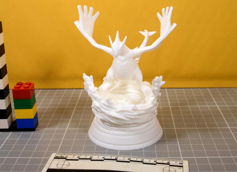 May include: A white 3D printed figurine of a mythical creature with large wings and a tail, standing on a sculpted base resembling swirling water. A ruler is visible at the bottom, showing the base width is approximately 3.5 inches wide.