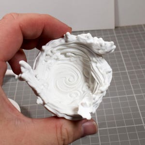 May include: A white 3D printed bowl featuring a sculpted, swirling water wave design that rises along the interior and exterior edges. The surface texture mimics the appearance of fluid motion with raised crests and a central spiral pattern.