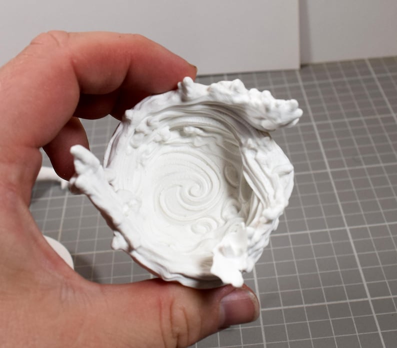 May include: A white 3D printed bowl featuring a sculpted, swirling water wave design that rises along the interior and exterior edges. The surface texture mimics the appearance of fluid motion with raised crests and a central spiral pattern.