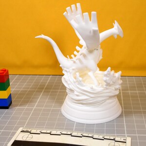 May include: A white 3D printed figurine of a winged creature with a long tail, positioned on a base resembling splashing water. A plastic ruler is visible at the bottom, showing a scale in centimeters, next to a stack of four colorful building blocks.