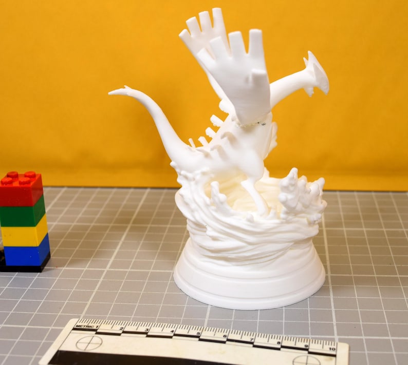 May include: A white 3D printed figurine of a winged creature with a long tail, positioned on a base resembling splashing water. A plastic ruler is visible at the bottom, showing a scale in centimeters, next to a stack of four colorful building blocks.