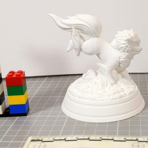 May include: A white, unpainted 3D printed figurine of a mythical creature with a flowing tail and mane, mounted on a circular base. The figure is positioned next to a stack of colorful plastic building blocks and a ruler for scale reference.