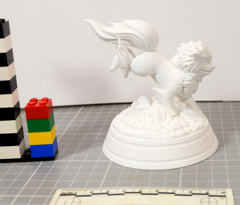 May include: A white, unpainted 3D printed figurine of a mythical creature with a flowing tail and mane, mounted on a circular base. The figure is positioned next to a stack of colorful plastic building blocks and a ruler for scale reference.