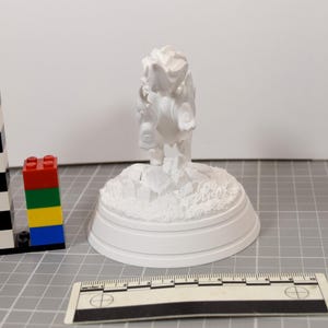 May include: A white 3D printed figurine of a creature standing on a rocky base, positioned next to a black and white plastic building block tower and a ruler showing measurements in centimeters, with the figurine measuring approximately 10 cm tall.