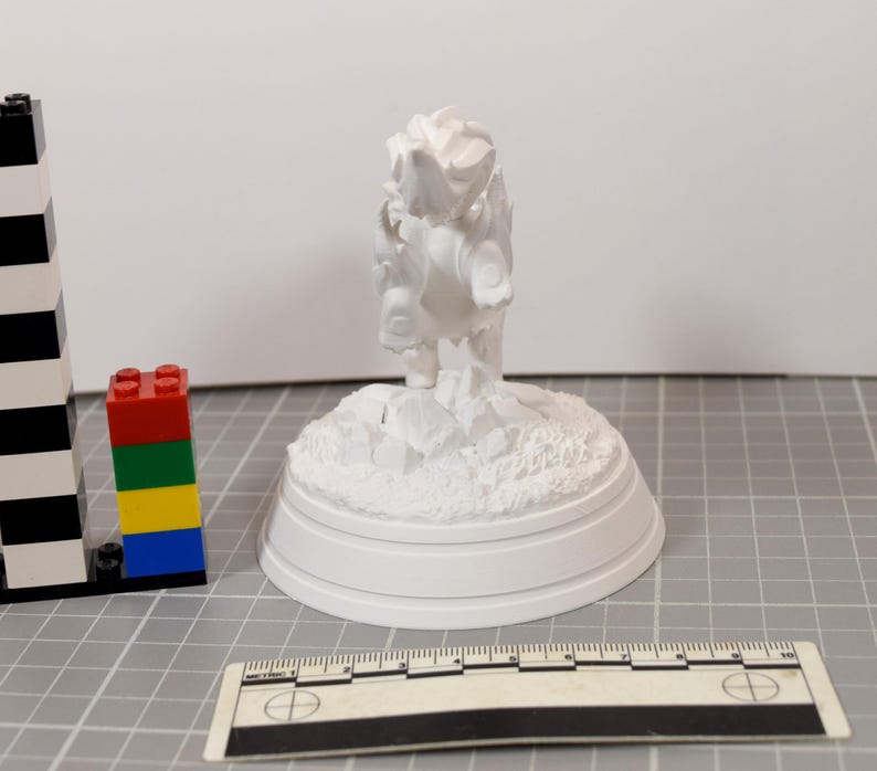 May include: A white 3D printed figurine of a creature standing on a rocky base, positioned next to a black and white plastic building block tower and a ruler showing measurements in centimeters, with the figurine measuring approximately 10 cm tall.