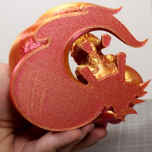 May include: A 3D printed figurine featuring a metallic gold creature with intricate embossed details, framed by a large, textured, glittery red crescent-shaped element that curves around the central figure with sharp, pointed edges.