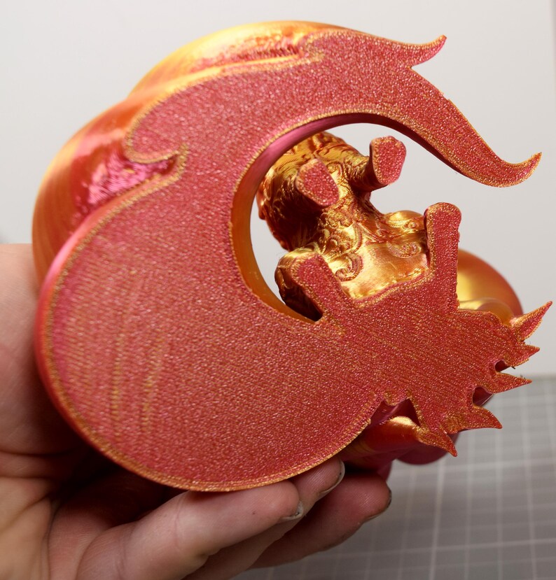 May include: A 3D printed figurine featuring a metallic gold creature with intricate embossed details, framed by a large, textured, glittery red crescent-shaped element that curves around the central figure with sharp, pointed edges.