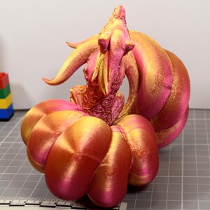 May include: A 3D printed figurine featuring a creature with a large, rounded, segmented base and a central upright form with intricate textures. The object displays a gradient of metallic pink and gold colors, positioned next to a ruler.