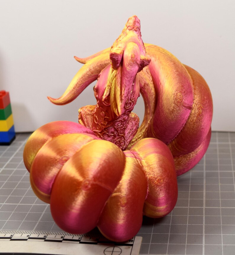 May include: A 3D printed figurine featuring a creature with a large, rounded, segmented base and a central upright form with intricate textures. The object displays a gradient of metallic pink and gold colors, positioned next to a ruler.