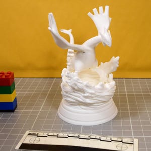 May include: A white 3D printed figurine of a winged creature emerging from a sculpted wave base. The figure is positioned next to a metric ruler and a small stack of red, green, yellow, and blue plastic building blocks on a grid surface.
