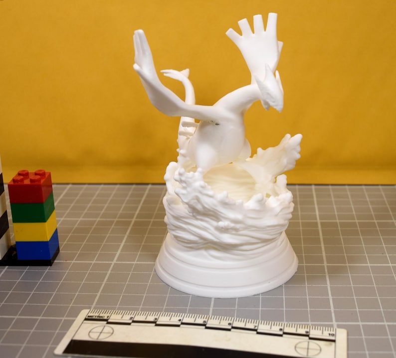 May include: A white 3D printed figurine of a winged creature emerging from a sculpted wave base. The figure is positioned next to a metric ruler and a small stack of red, green, yellow, and blue plastic building blocks on a grid surface.