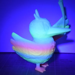 May include: A brightly coloured duck figurine with a rainbow gradient on its wings and chest. The duck has a long beak and is holding a long, thin object. The figurine is illuminated with a blue light, giving it a glowing appearance.