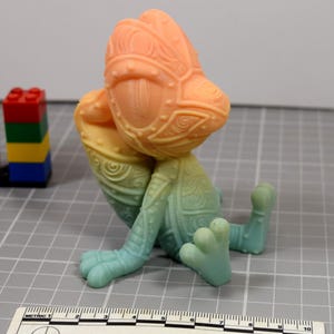 May include: A colorful, stylized chameleon figurine in shades of orange, yellow, and teal. The figurine has detailed patterns and sits with its legs extended. A stack of colorful toy blocks is in the background.
