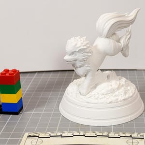 May include: A white, unpainted 3D printed figurine of a four-legged creature with a large, flowing tail and a textured mane, mounted on a circular base. A ruler is visible at the bottom, showing a scale in centimeters for size reference.