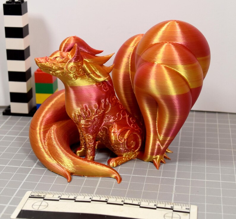 May include: A 3D printed fox figurine with multiple tails, featuring a gradient of metallic red, orange, and gold colors. The surface is adorned with intricate embossed scrollwork patterns. A ruler is visible in the foreground for scale.