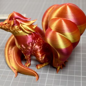 May include: A 3D printed fox figurine with multiple tails, featuring a gradient of metallic red, orange, and gold colors. The surface displays visible horizontal layer lines from the printing process, with intricate embossed patterns on the head and body.