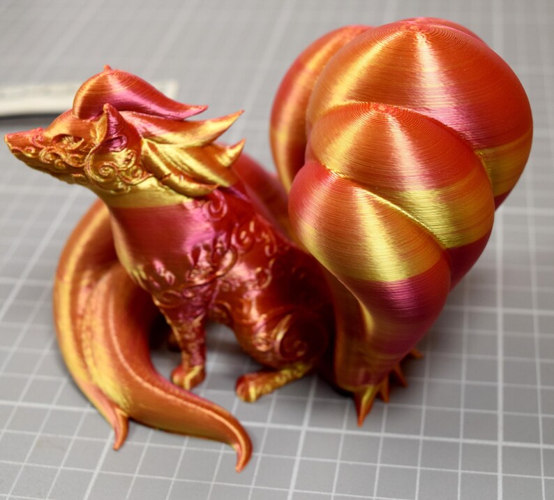 May include: A 3D printed fox figurine with multiple tails, featuring a gradient of metallic red, orange, and gold colors. The surface displays visible horizontal layer lines from the printing process, with intricate embossed patterns on the head and body.